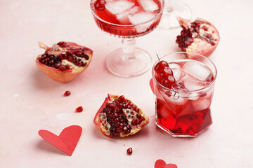 glasses with fresh pomegranate juice with one real pomegranate on concrete tray on pink background with red hearts decoration