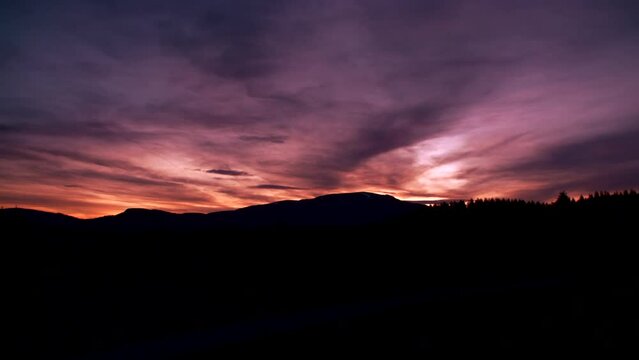 Stunning panning shot over the silhouette of forest and mountains against a gorgeous red purple sunset sky.