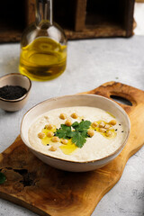 Hummus dip with chickpea and parsley in ceramic bowl on concrete surface with toasted baguette slices and olive oil on wooden board