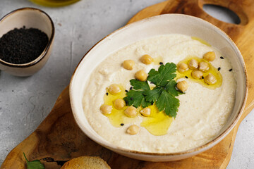 Hummus dip with chickpea and parsley in ceramic bowl on concrete surface with toasted baguette slices and olive oil on wooden board