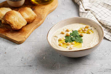 Hummus dip with chickpea and parsley in ceramic bowl on concrete surface with toasted baguette slices and olive oil on wooden board