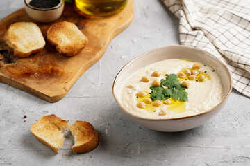Hummus dip with chickpea and parsley in ceramic bowl on concrete surface with toasted baguette slices and olive oil on wooden board