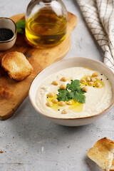 Hummus dip with chickpea and parsley in ceramic bowl on concrete surface with toasted baguette slices and olive oil on wooden board