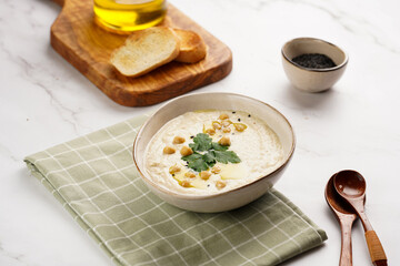Hummus dip with chickpea and parsley in ceramic bowl on marble surface with toasted baguette slices and olive oil on wooden board