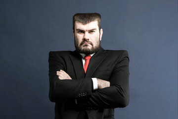 Boss. Confident young man. Businessman. The leader looks at the subordinate. A brunette man with a beard in a business suit with a red tie on a blue background crossed his arms. Politician