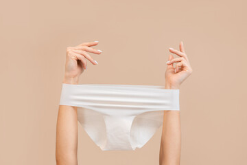 Elegant female hands with panties stretched over them. Beige background. Copy space. The concept of gynecology and depilation - obrazy, fototapety, plakaty