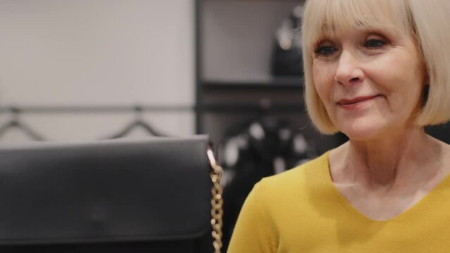 Mature woman middle-aged client in store female accessories chooses goods searching for suitable purchase surprised rejoices looking at appropriate ladies handbag smiling happy buying at discount sale