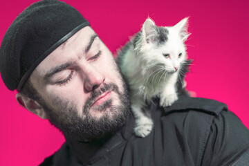 A male security guard in a black uniform and a cap holds a cute fluffy kitten on his shoulder. The...