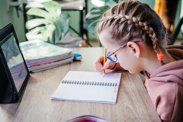 Primary school girl in eyeglasses writing in her notebook while having online lesson during covid...