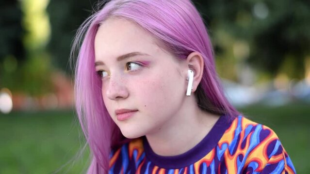 Pink-haired teenage hipster girl in a colorful bright T-shirt and earphones is using a smartphone on a city street on a summer day. Summer concept. Generation Z style. Social media concept. Slow motio