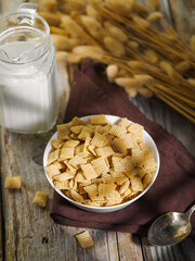 On a brown napkin, a bowl of crispy cereal snacks, a bunch of cereals and fresh milk in a jug. Wooden texture. Quick breakfasts, minerals, vitamins, carbohydrates, fiber. Healthy lifestyle.