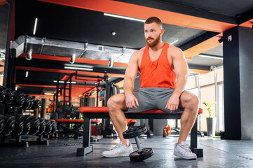 Fototapeta na wymiar A young bearded athletic man is sitting on a sports bench. The dumbbell lies near the athlete's feet. Bodybuilding and fitness concept