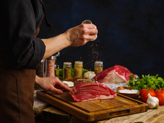 Cooking juicy beef steak by chef's hands on a wooden table, dark blue background. Lots of spices, herbs, vegetables. Restaurant, hotel, cafe, food blog.