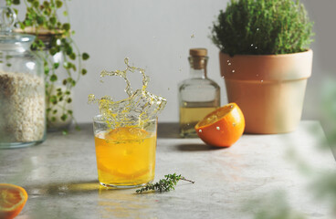 Splash of orange lemonade with herbs at kitchen table with glass jar, bottle, halved orange and potted kitchen herbs at wall . Refreshing summer drink. Front view.