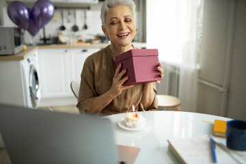 Happy mature woman of 50s smiling shaking present box to guess what's inside sitting in front of laptop and cupcake with candle against balloons in her kitchen, celebrating birthday online