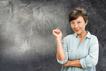 Photo of beautiful old Asian woman with gray hair, wearing glasses and blue shirt, standing on dark gray concrete wall background. Copy space backdrop, artwork. Elegant happy elderly retire concept