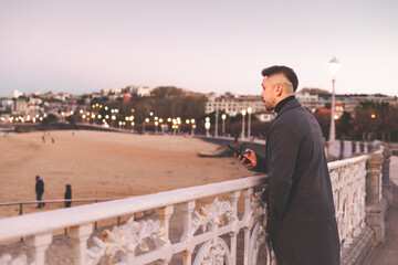 Fototapeta na wymiar Young caucasian man using a smartphone at Donostia-San Sebastian sea walk during sunset time.