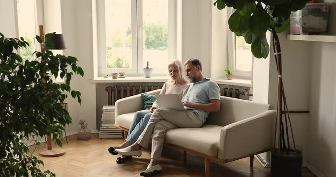 In cozy light living room senior spouses sit on sofa using laptop, make order through distancing electronic online services. Older generation and modern wireless tech usage, e-commerce clients concept