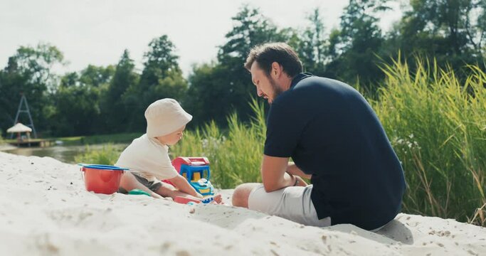 Dad spending time with his son in the midst of nature, they are sitting on the beach near the lake, surrounding greenery, playing in the sand, toys around, shovel, rakes, bucket