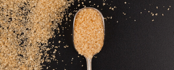 A teaspoon filled with brown sugar, lying on a black tabletop with sugar spilled over it.