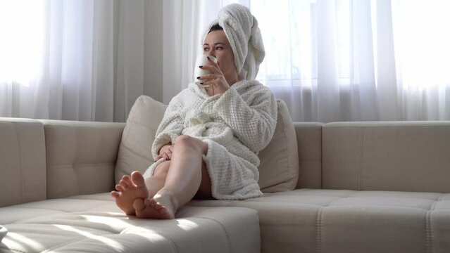 Portrait of a beautiful girl, after a shower in a white bathrobe and with a towel on her head, drinks tea on the sofa, stretching her legs and looking out the window, medium plan, soft focus