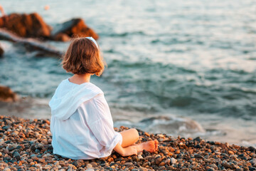 Summertime. Pre-school girl sitting alone on the pebble beach. Back view. Ocean at the background. Concept of adoption and children's day