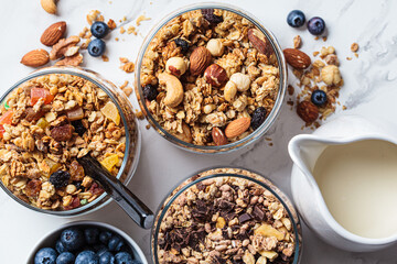 Different types of homemade granola (fruit, nut and chocolate) in jars on white kitchen table. Breakfast food concept.