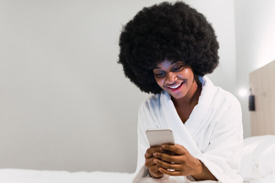 Afro woman smiling and using mobile phone on bed at home