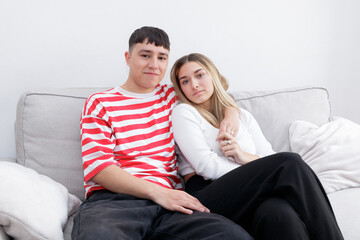 Happy embraced boy and girl sitting in the sofa smiling looking at camera. Young couple at home. Heterosexual 18-20 years old couple.