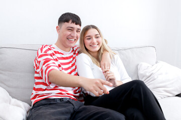 Boyfriend and girlfriend watching TV laughing. Boy pointing to the screen. Young couple at home. Heterosexual 18-20 years old couple.