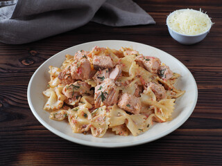 Pasta farfalle with pieces of fish salmon in cream sauce with parmesan cheese in a plate, napkin on a dark table, flat layout. Traditional delicious dish with curly  pasta products and seafood