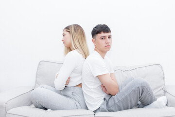 Angry couple sitting in the sofa back to back with crossed arms. Man looking at camera. Mad couple after a relationship conflict.  Young couple at home. Heterosexual 18-20 years old couple.