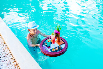 cute school boy with served floating tray in swimming pool with drinks and snacks on tropical...
