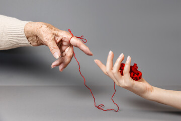Hands of a young and an senior woman reach out to each other, connected by a red thread. Gray...