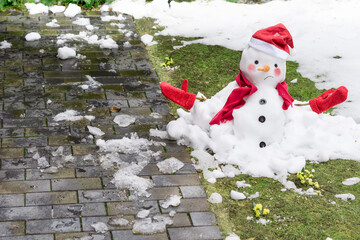 Unhappy snowman in mittens, red scarf and cap is melting  outdoors in sunlight on snowy green grass...