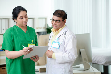 Focused mature male doctor and female nurse looking at screen disc of tablet computer and discussing anamnesis of patient - obrazy, fototapety, plakaty