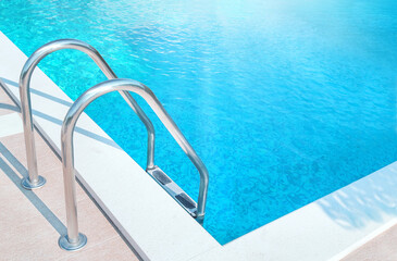 Ladder stainless handrails (stairs) into the swimming pool with blue water and sunlight (blik of...