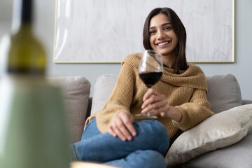 Pretty, attractive, stylish young woman sitting on couch having raised glass with red wine, examine, taste beverage