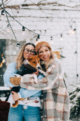 Portrait of two cheerful women friends standing outdoor in the backyard with dog in snowfall, enjoying winter time.