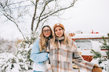 Portrait of two cheerful women friends standing outdoor near van during the snowfall in winter camp.