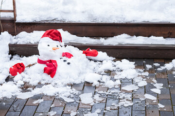 Unhappy snowman in knitted mittens, red scarf and cap is melting  outdoors, on pavement,  snowy background. Approaching spring, warm winter, climate change concept  