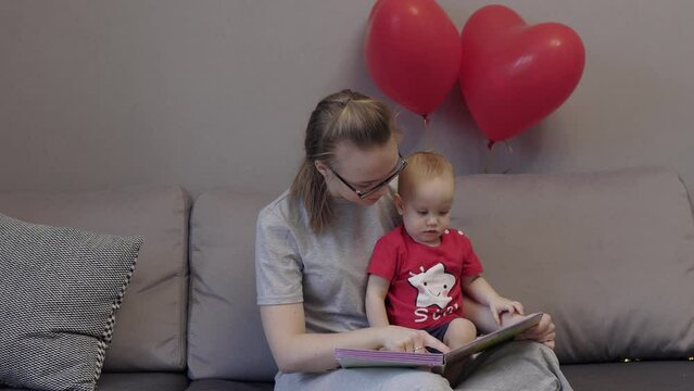 young caucasian mother in glasses reading education book to her baby child kid in red t-shirt. mother and toddler sitting grey sofa in living room looking picture book with red heart balloons behind