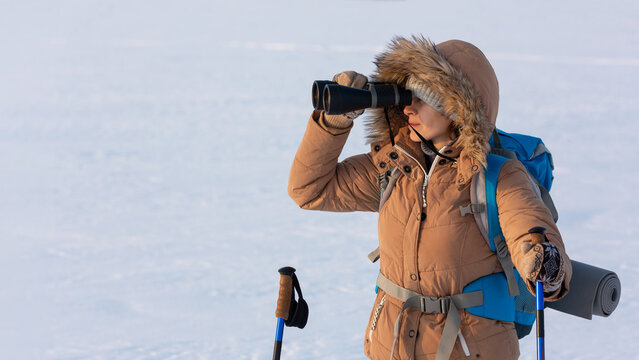 Woman in yellow hooded down jacket with a backpack and ski poles looks through binoculars standing on a snowy plain in winter, waist up, selected focus.