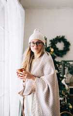 Young caucasian woman standing near window in the morning, drinking tea and enjoying winter time.