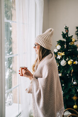 Young caucasian woman standing near window and looking outside in the morning, drinking tea and enjoying winter time.