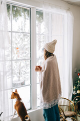 Young caucasian woman standing near window with her dog and looking outside in the morning, drinking tea and enjoying winter time.