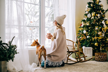 Young caucasian woman sitting near window with her dog and looking outside in the morning, drinking...