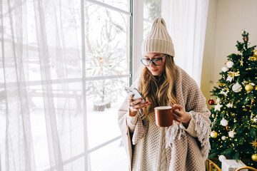 Young caucasian woman standing near window and using mobile phone, drinking tea in winter morning.