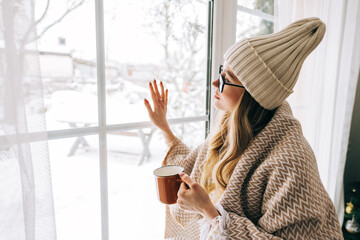 Young caucasian woman standing near window and looking outside in winter holidays, enjoying cozy atmosphere.