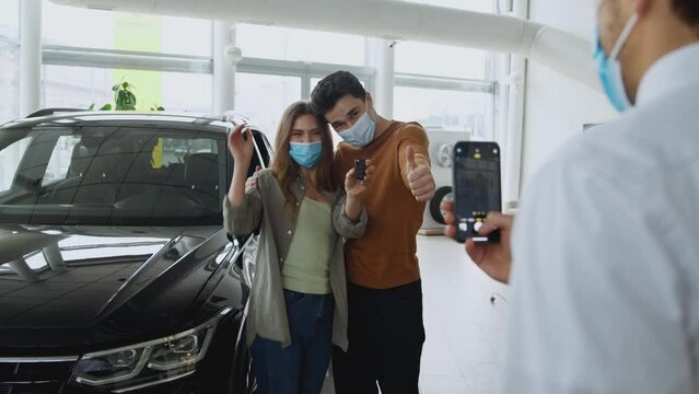 Young happy couple in love wearing face masks posing with new car and keys at showroom. salesman photographing them